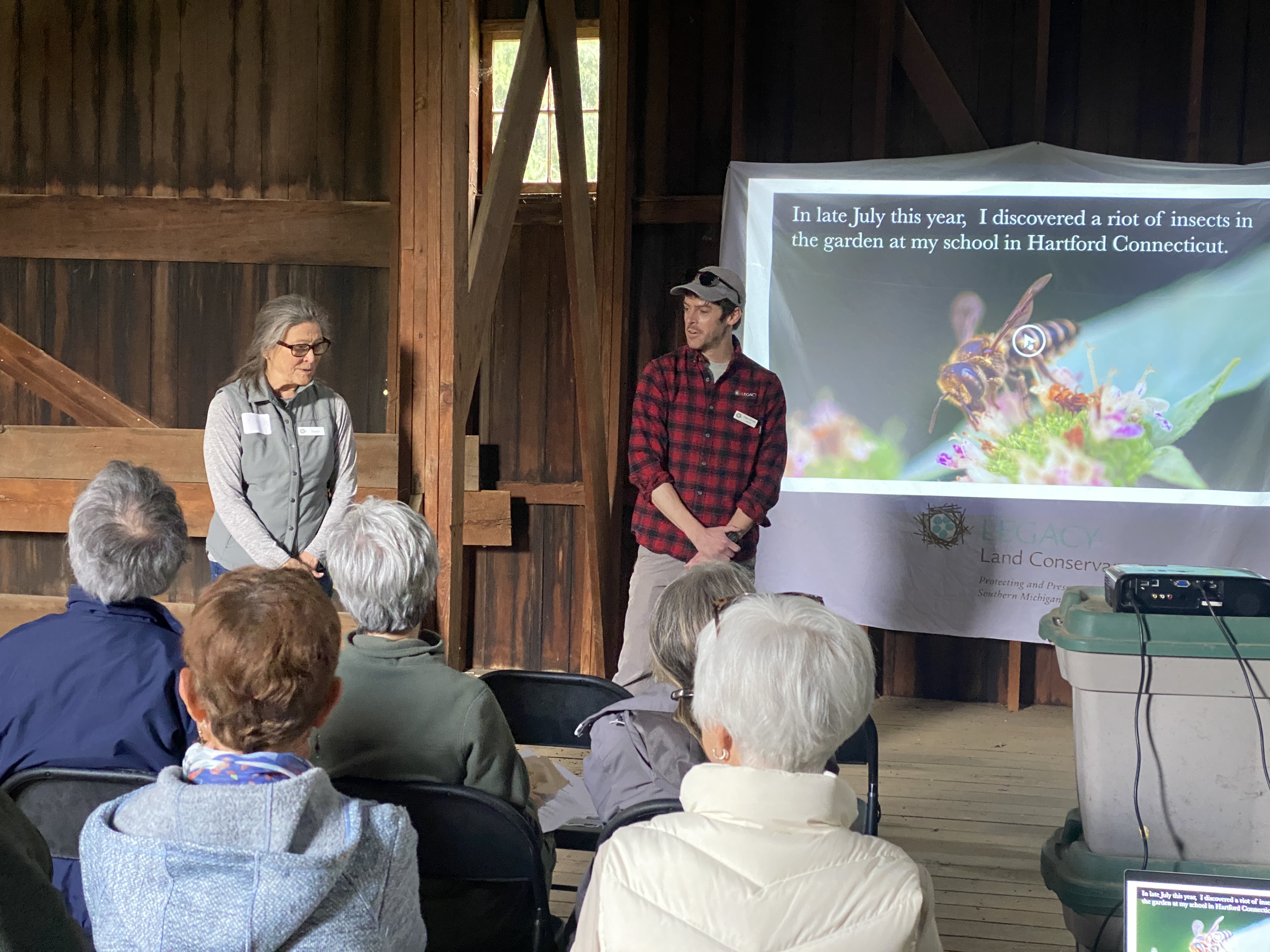 Two people standing in front of a large screen talking to a group of people sitting in a barn