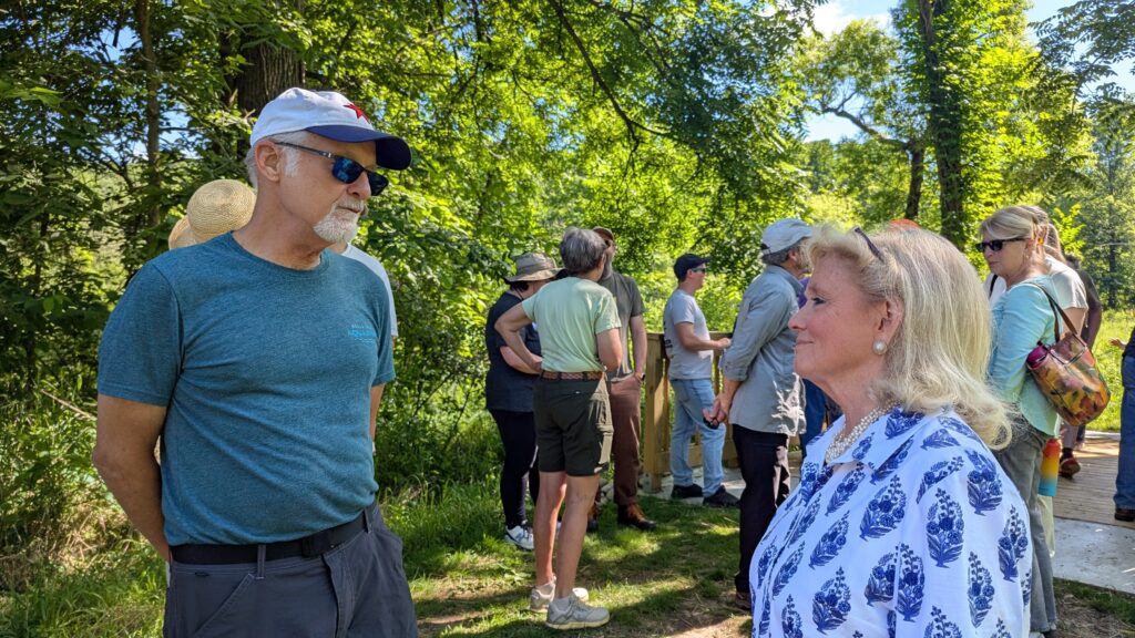 Two people smiling and talking. Many other people in the background with green trees behind them.