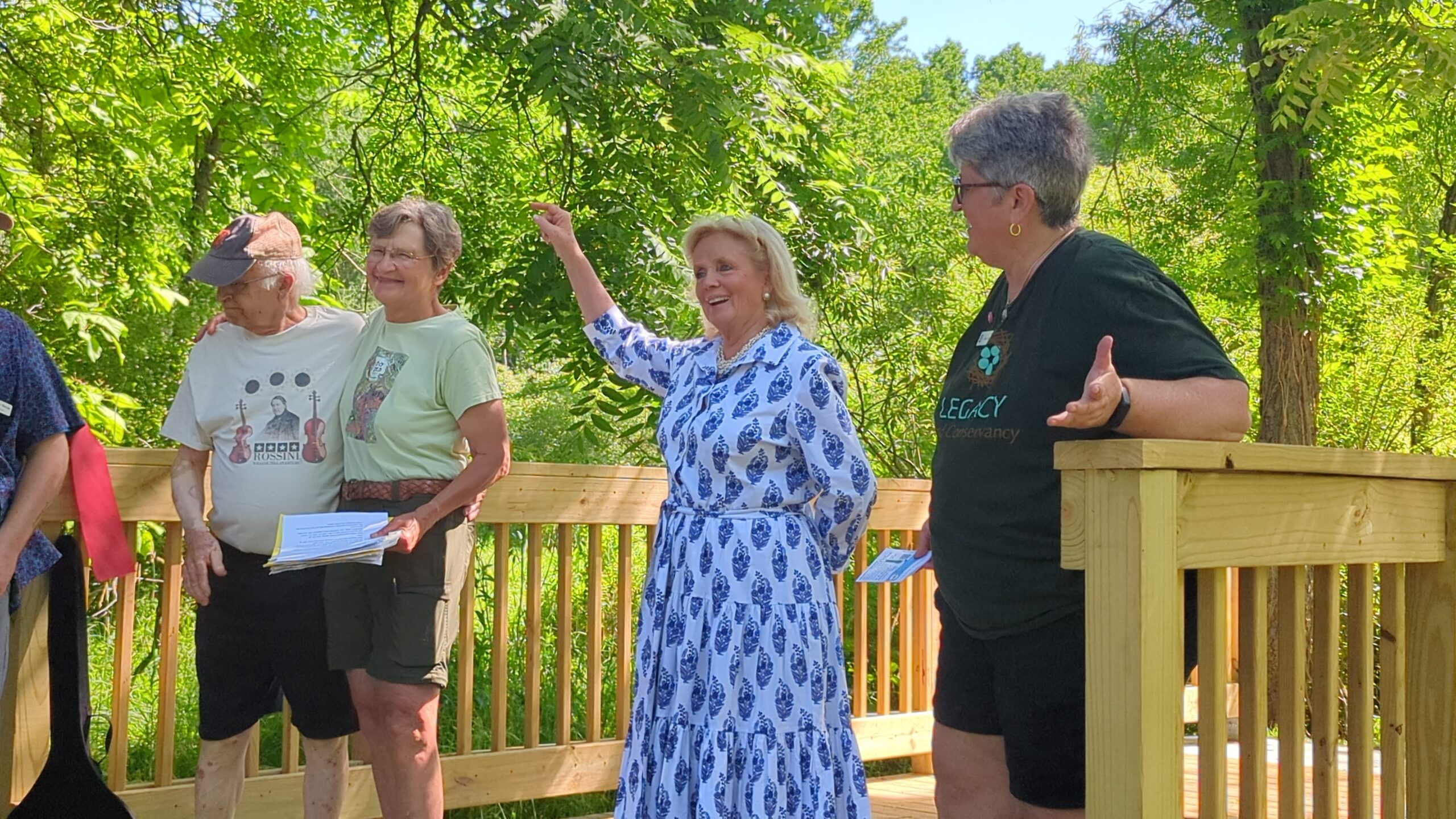 Four people standing on a wooden bridge. One in the middle holds their hand up and points at a couple smiling.