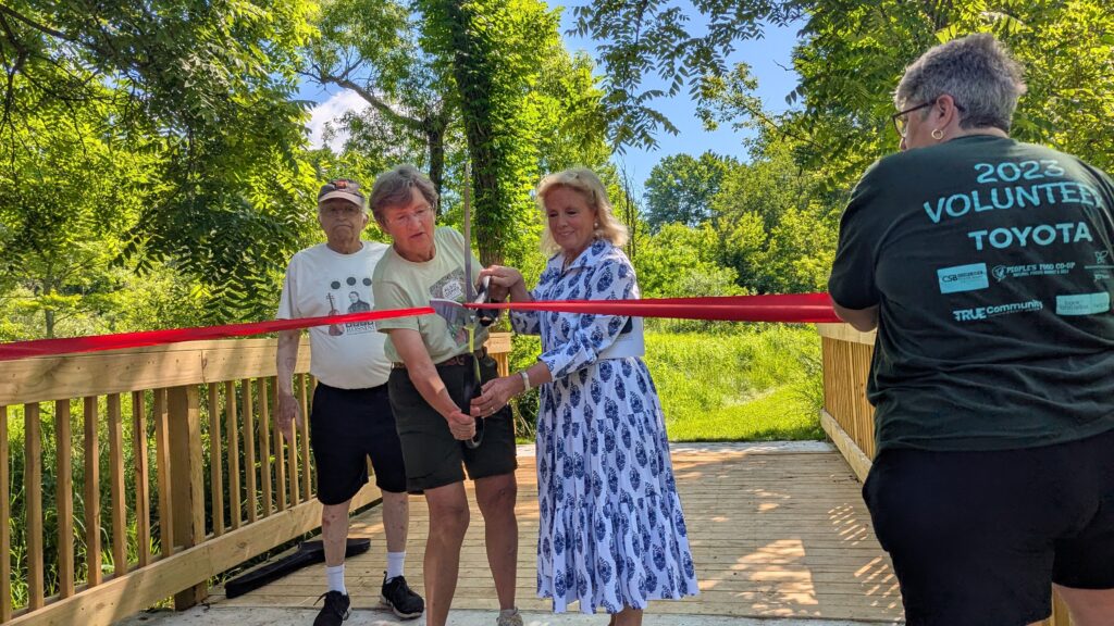 Four people stand on a bridge. Two people hold very large scissors on a big red ribbon.