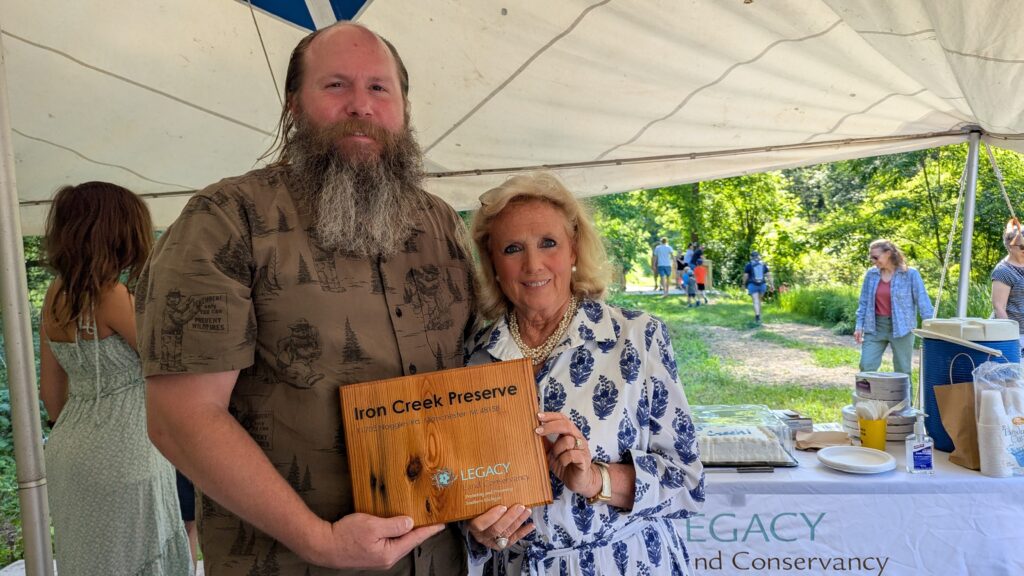 Two people pose for a picture while holding a wooden sign that says Iron Creek Preserve and Legacy Land Conservancy.