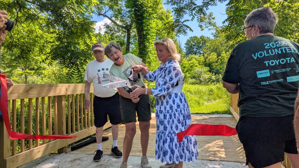 Four people on a wooden bridge. Two people use very large scissors to cut a big red ribbon.