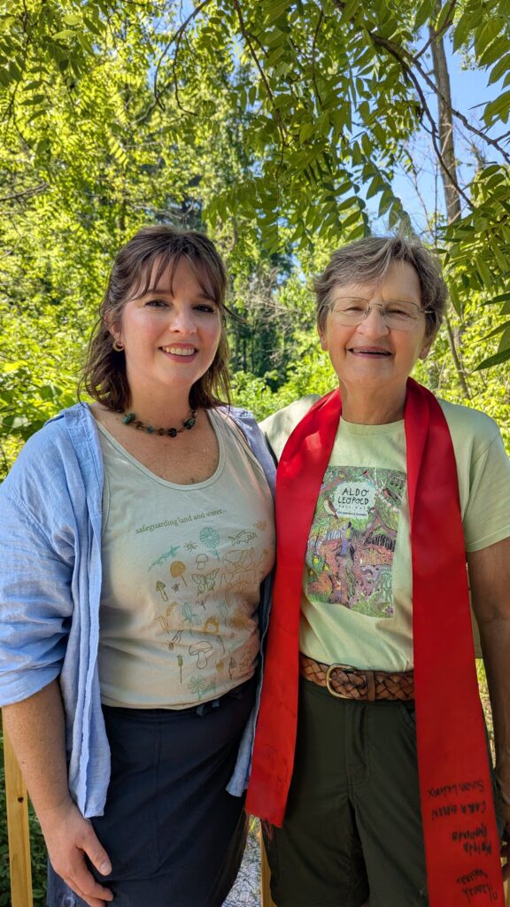 Two people smiling and standing with green trees and blue sky in the background.