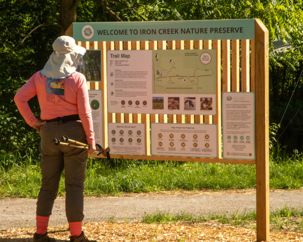 One person wearing a hat and holding walking sticks looks at a Welcome to Iron Creek Preserve kiosk sign.