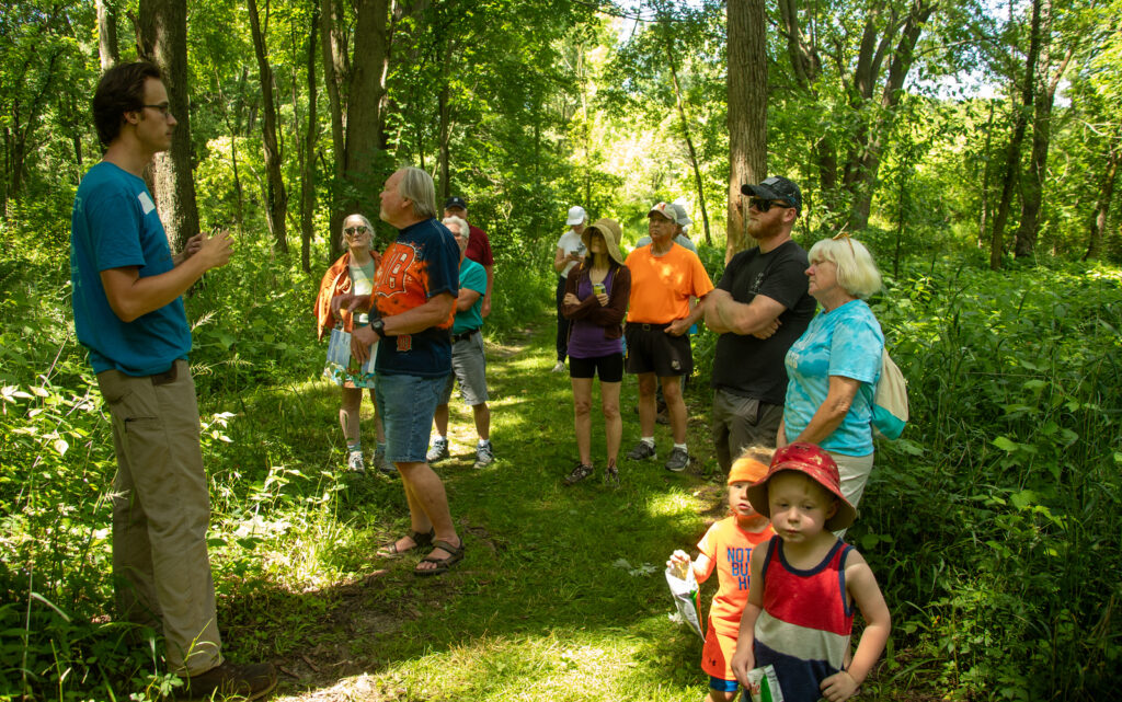 Many people standing on a green path listening to one person speak. Green trees and green plants surround them.