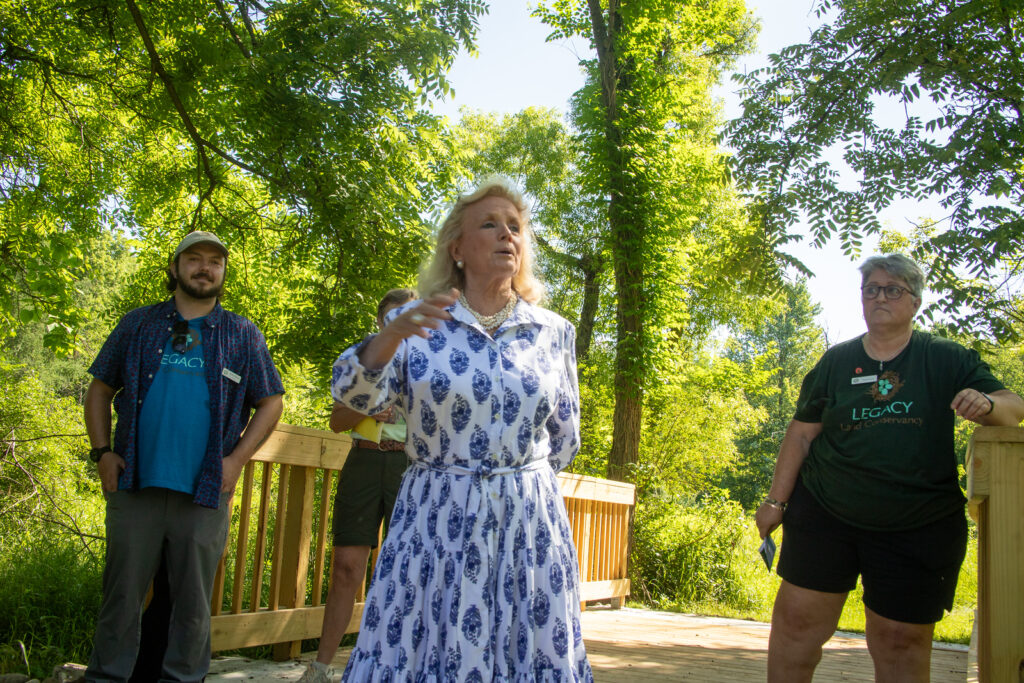 Four people stand on a bridge. One is speaking. Green trees and blue sky in the background.