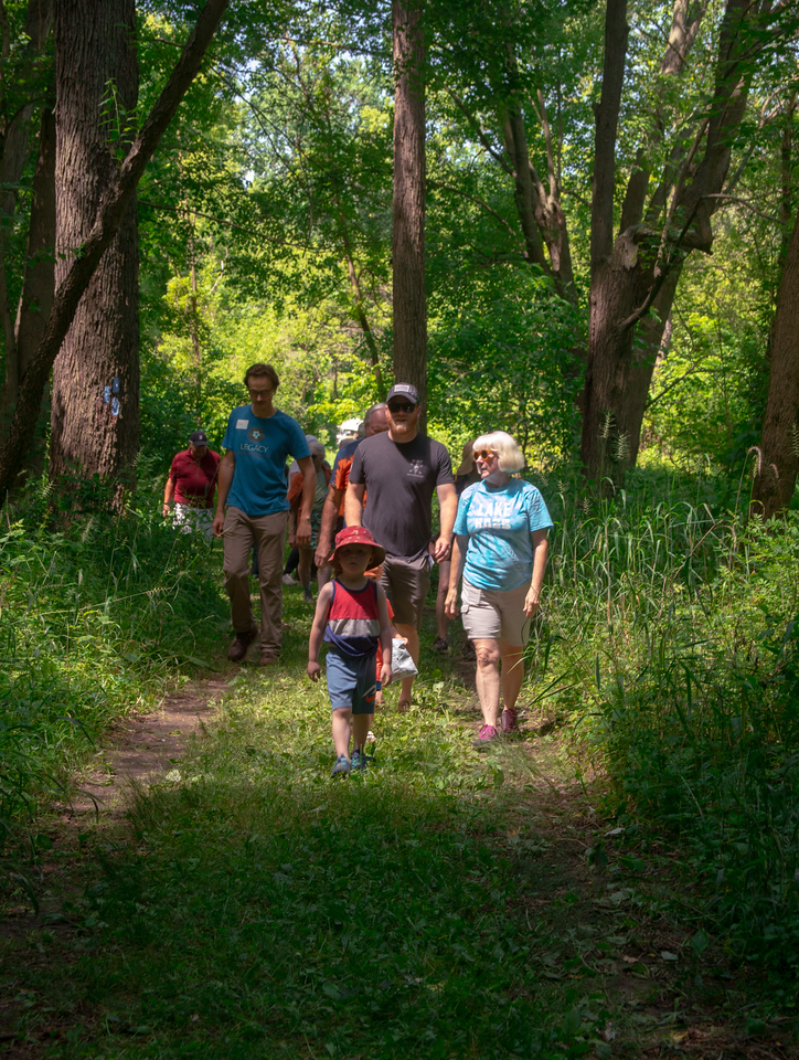 Kid and adults walking on a dirt and green path with tall green trees above them.