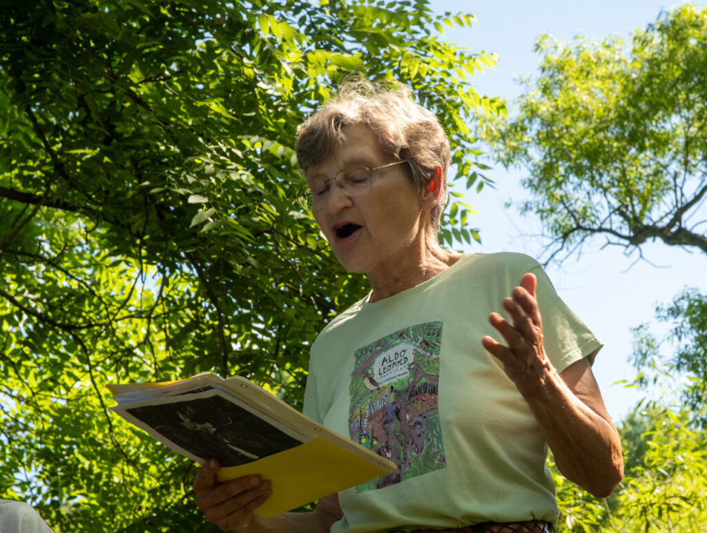 Person reading from a notepad with green trees and blue sky behind them.