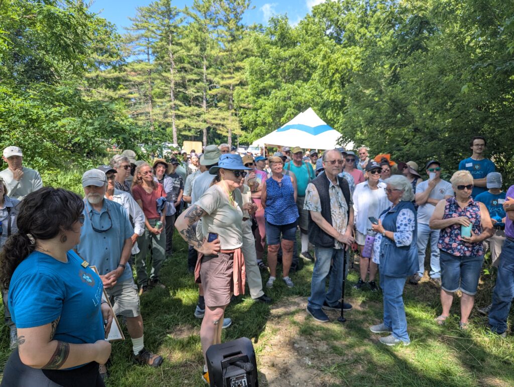 Many people stand in a narrow path. Green trees, a white tent, and blue sky in the background.
