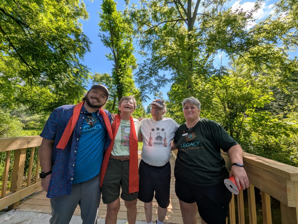 Four people smiling and standing on a wooden bridge with green trees and blue sky in the background.