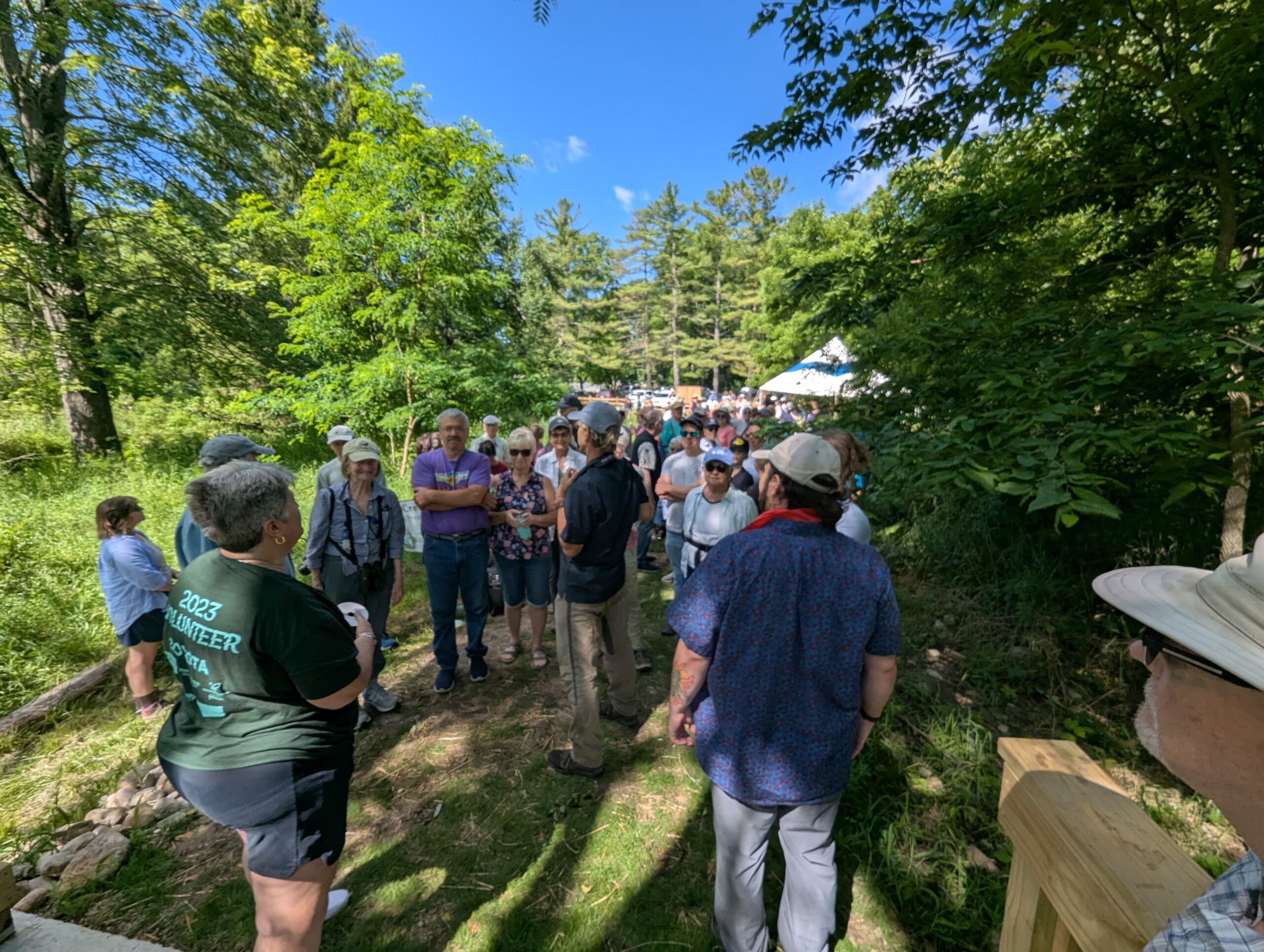 Many people stand huddled on a narrow path. Green trees, a white tent, and blue sky in the background.