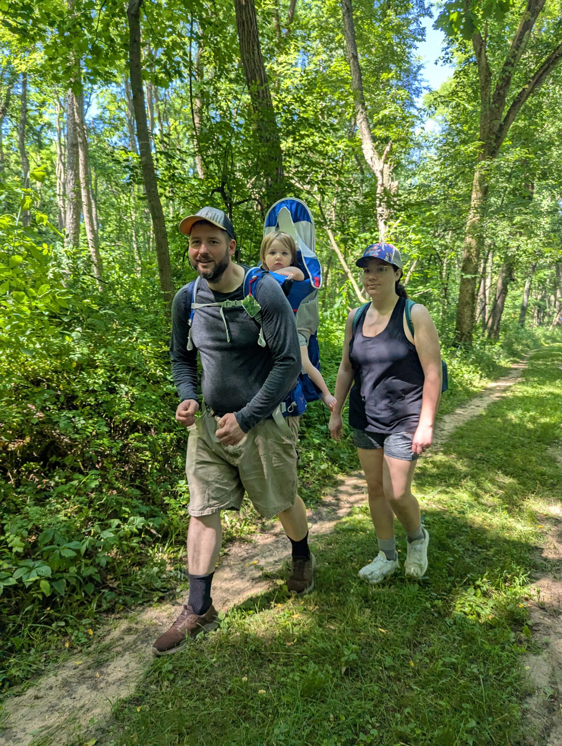 Two adults and one child in a backpack carrier walk on a green trail surrounded by green trees.
