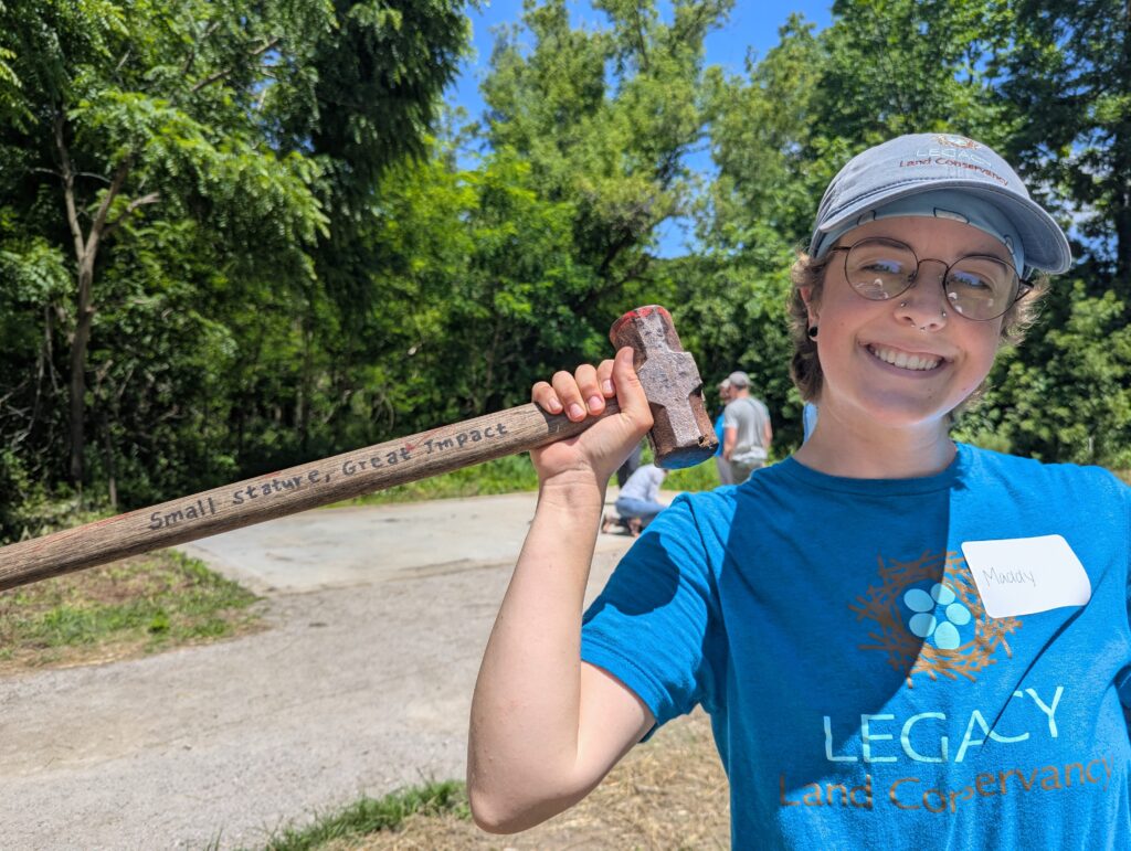 Person wearing Legacy Land Conservancy shirt and hat holds sledge hammer with the words "Small stature, Great impact" written on the wood handle