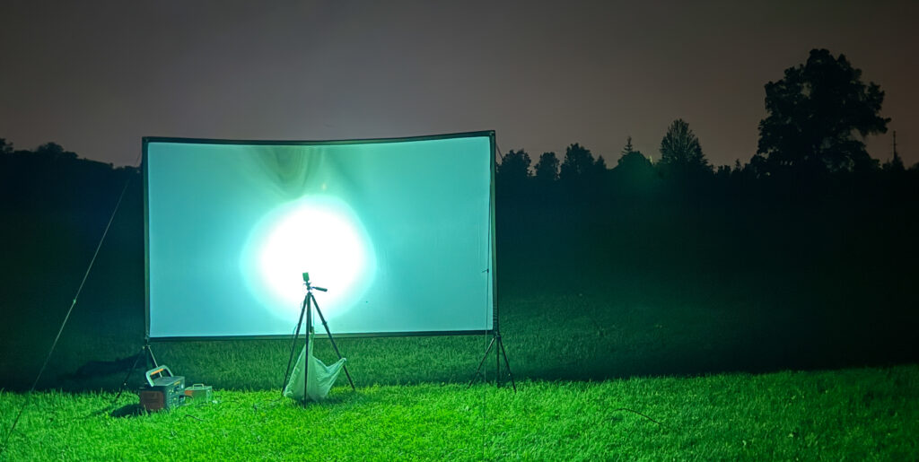 Large rectangular sheet on grassy field. Night sky and trees in the background. Bright light shines on the screen.