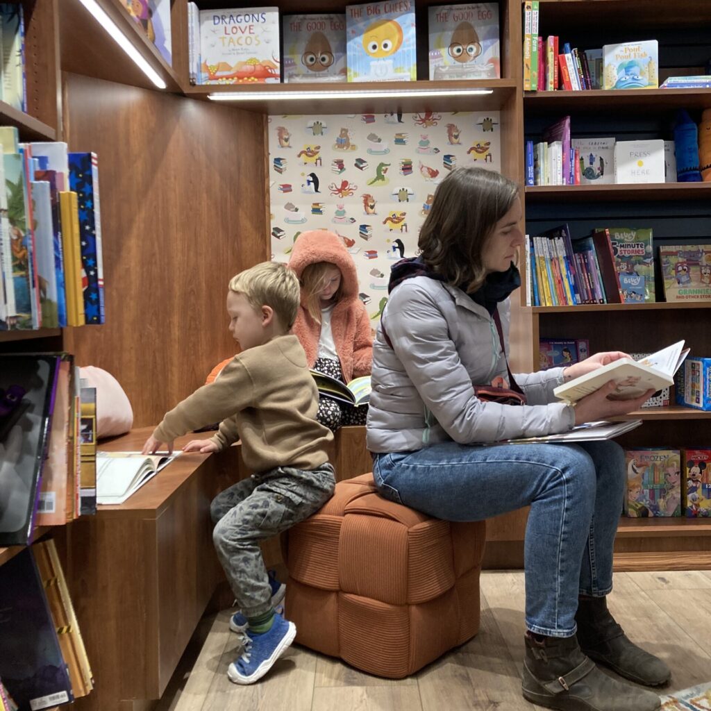 One adult and two kids sitting on a cushion reading books in a bookstore.