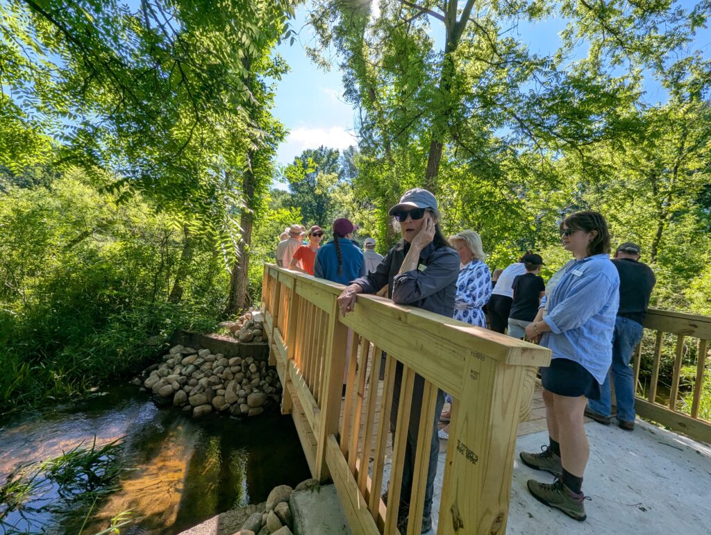 Adults stand on a wooden bridge over a small creek. Trees and blue sky in the background.
