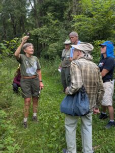 Person speaking to a small group of people with a forest of trees in the background.