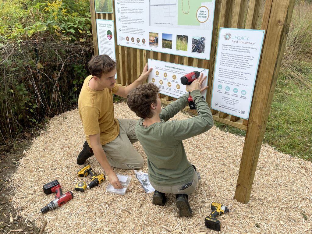 Two people screw in signs on a wooden kiosk.