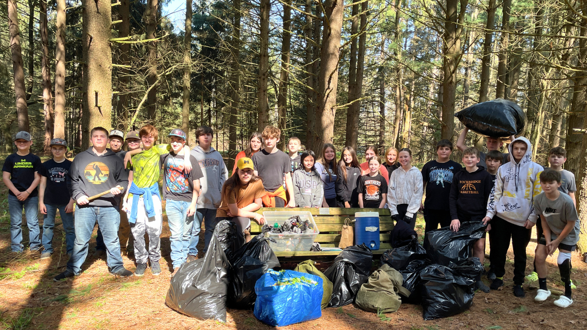 Many students stand in a forest of pine trees with garbage bags.
