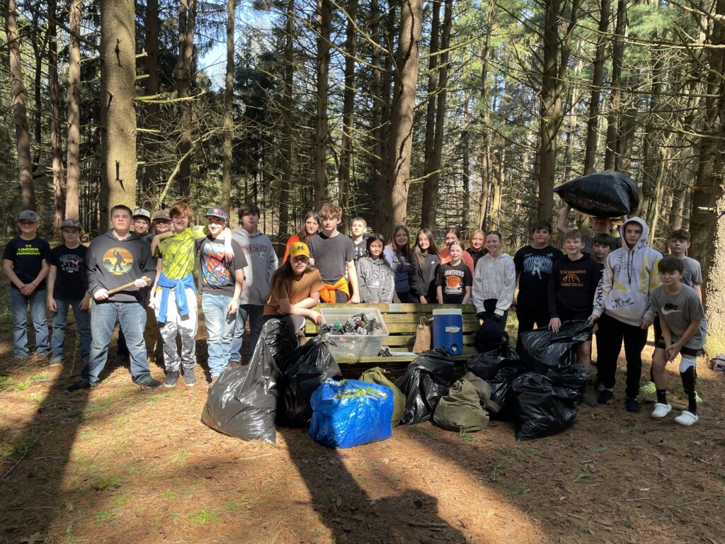 A large group of students stand in a forest next to garbage bags full of invasive plants.